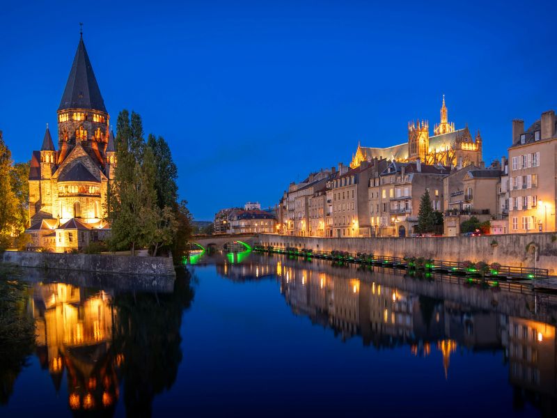 フランス・メス、ブルーアワーに照らされたモゼル川沿いの夜景。左手にテンプル・ヌフ、右奥にゴシック建築のメス大聖堂がライトアップされ、川面に光が映り込んでいる