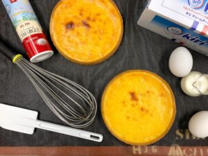 こんがり焼き色のついたチーズタルト2個と、フランス・カマルグ産海塩、キリ・クリームチーズ(ベル社)、卵、生クリーム、泡立て器、ゴムベラを黒いマットの上に並べた製菓風景の写真